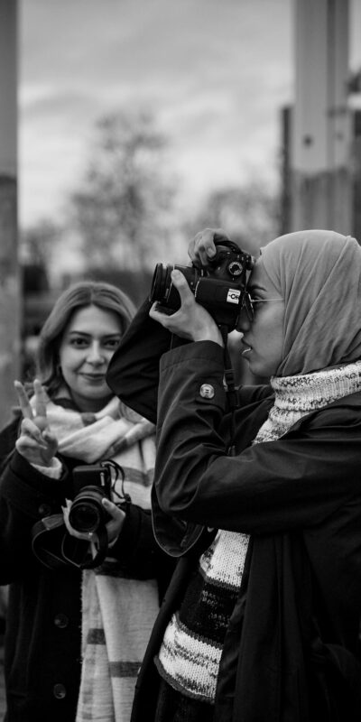 Black and white photo of female photographers outdoors, one taking a picture and another posing with a peace sign.